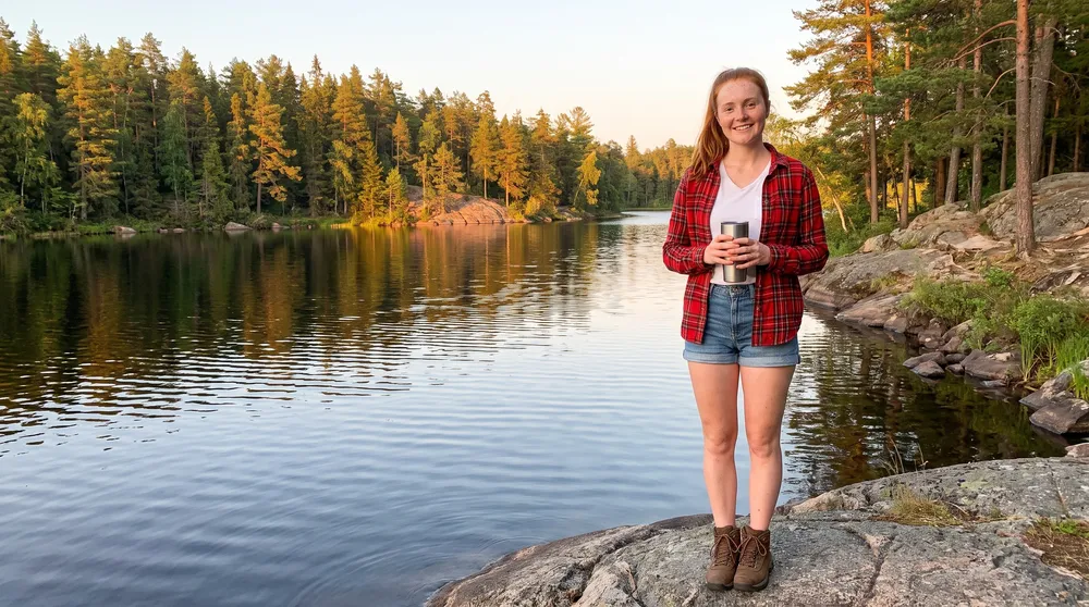 Ellie Marchand stands on a granite outcrop beside a calm lake ringed by spruce, holding a steel travel mug.